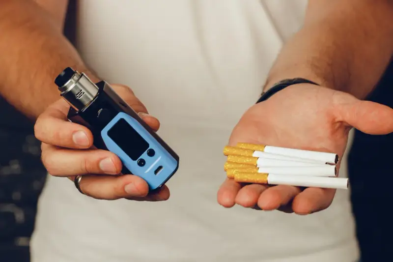 Man holds a vape in one hand and cigarettes in the other, posing the question: que es mejor el vaper o el tabaco.
